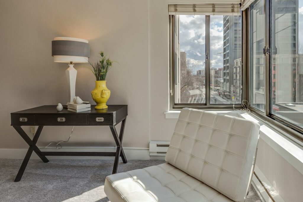 Coin salon lumineux avec un fauteuil blanc, une lampe et un vase jaune près de grandes fenêtres.