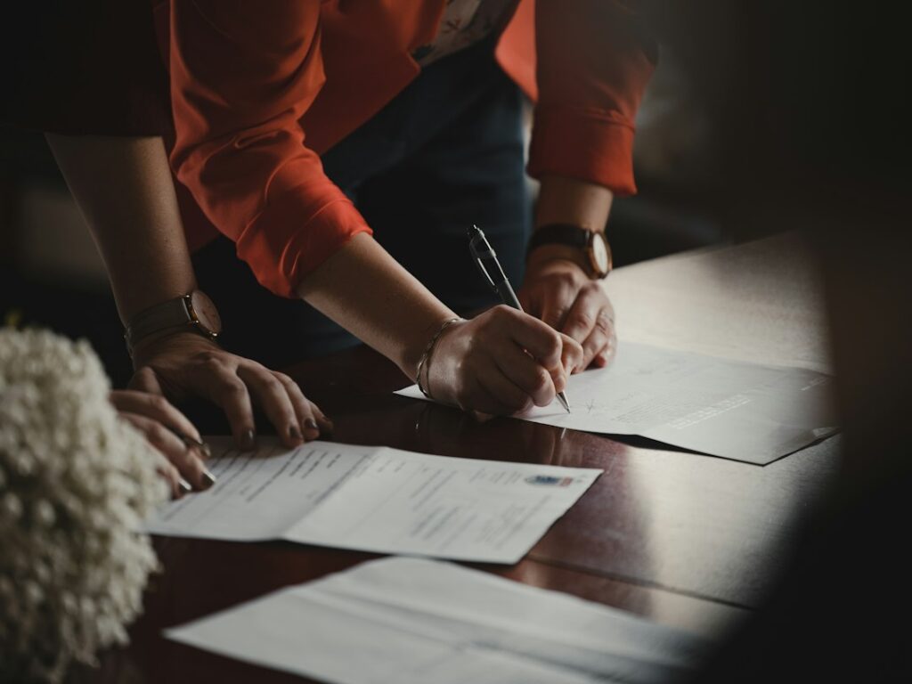 Mains de deux personnes autour de documents, dont l’une écrit avec un stylo sur une table en bois.