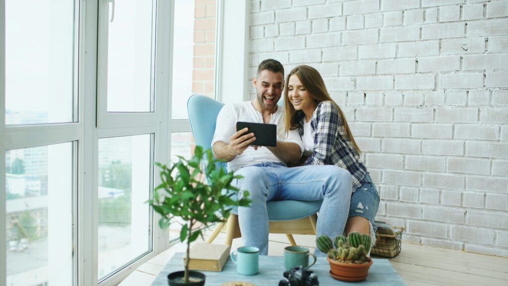Un couple consulte une tablette près d’une grande fenêtre, dans un salon aux tons blancs et bleus.