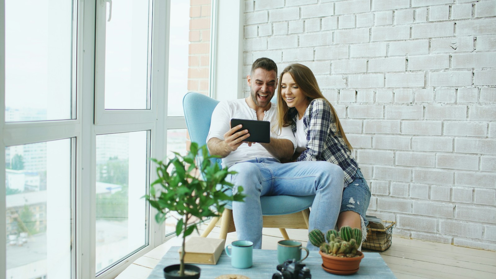 Un couple consulte une tablette près d’une grande fenêtre, dans un salon aux tons blancs et bleus.